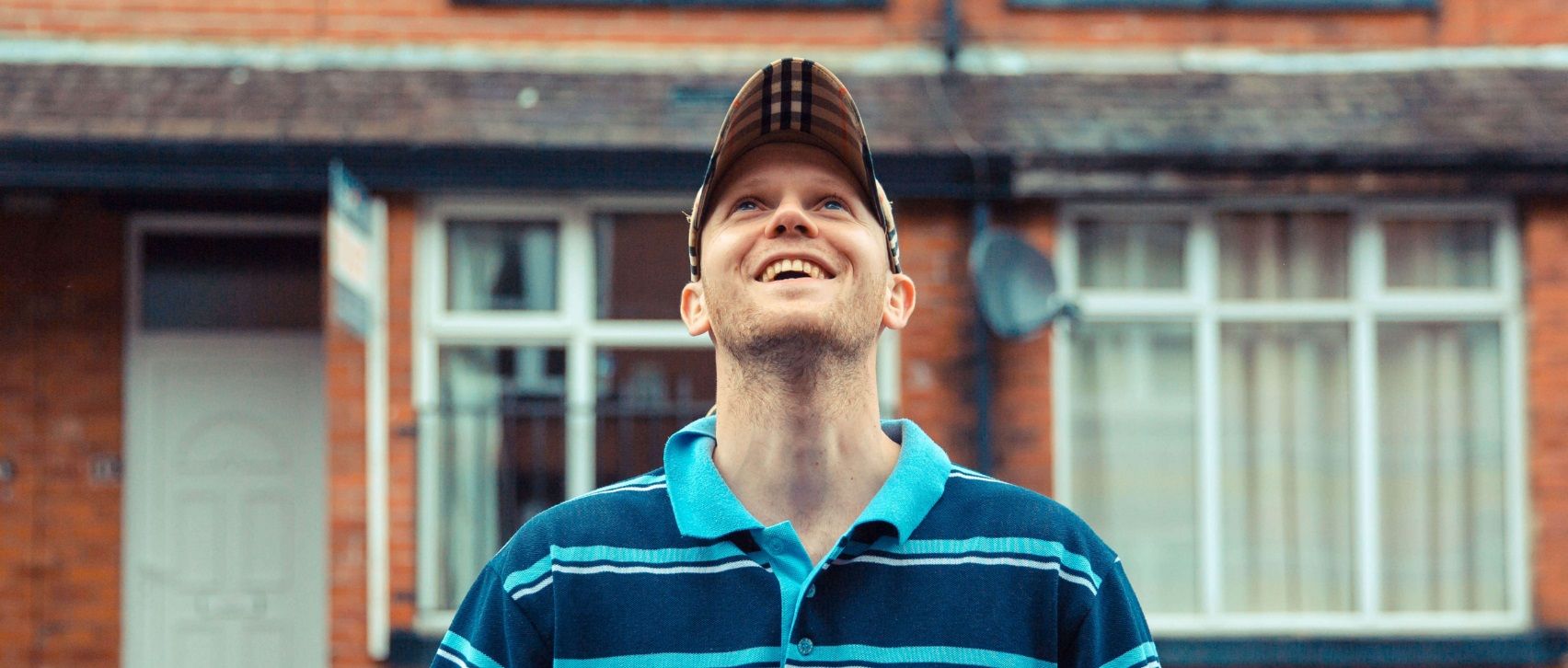 Young man wearing a baseball cap and striped polo shirt smiling and looking up while standing in front of brick terraced houses.