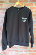 A black long-sleeved crewneck sweatshirt hanging on a wooden hanger against a red brick wall. The left chest features a white "Devonshire Trading Company" bird logo.