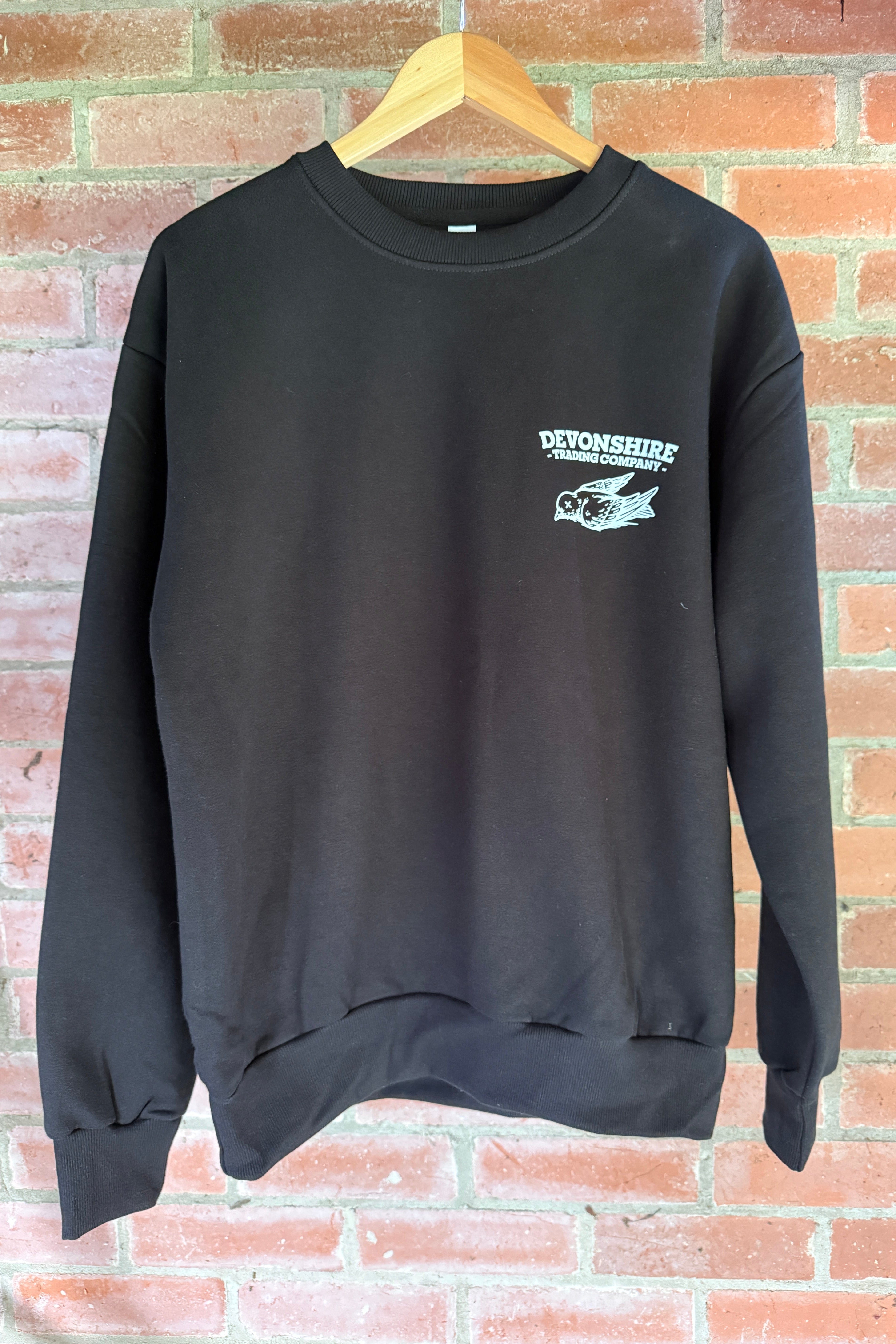 A black long-sleeved crewneck sweatshirt hanging on a wooden hanger against a red brick wall. The left chest features a white "Devonshire Trading Company" bird logo.