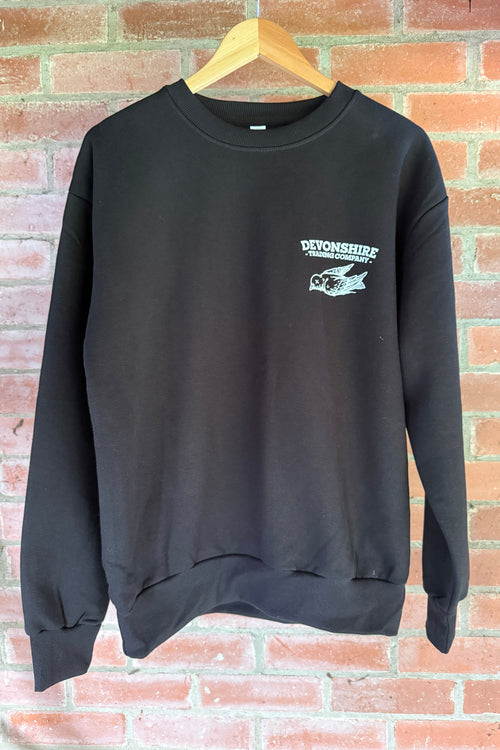 A black long-sleeved crewneck sweatshirt hanging on a wooden hanger against a red brick wall. The left chest features a white "Devonshire Trading Company" bird logo.