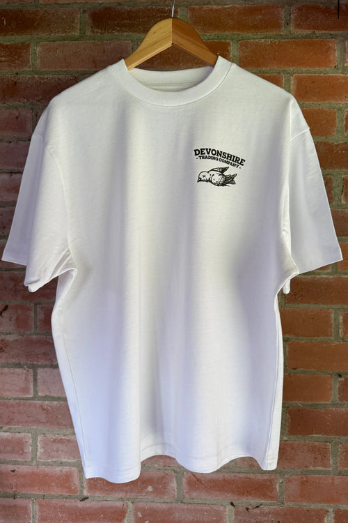 A white short-sleeved t-shirt hanging on a wooden hanger against a red brick wall. The shirt features a small black graphic on the left chest area: a stylized bird illustration with the text "DEVONSHIRE TRADING COMPANY" curved above it.